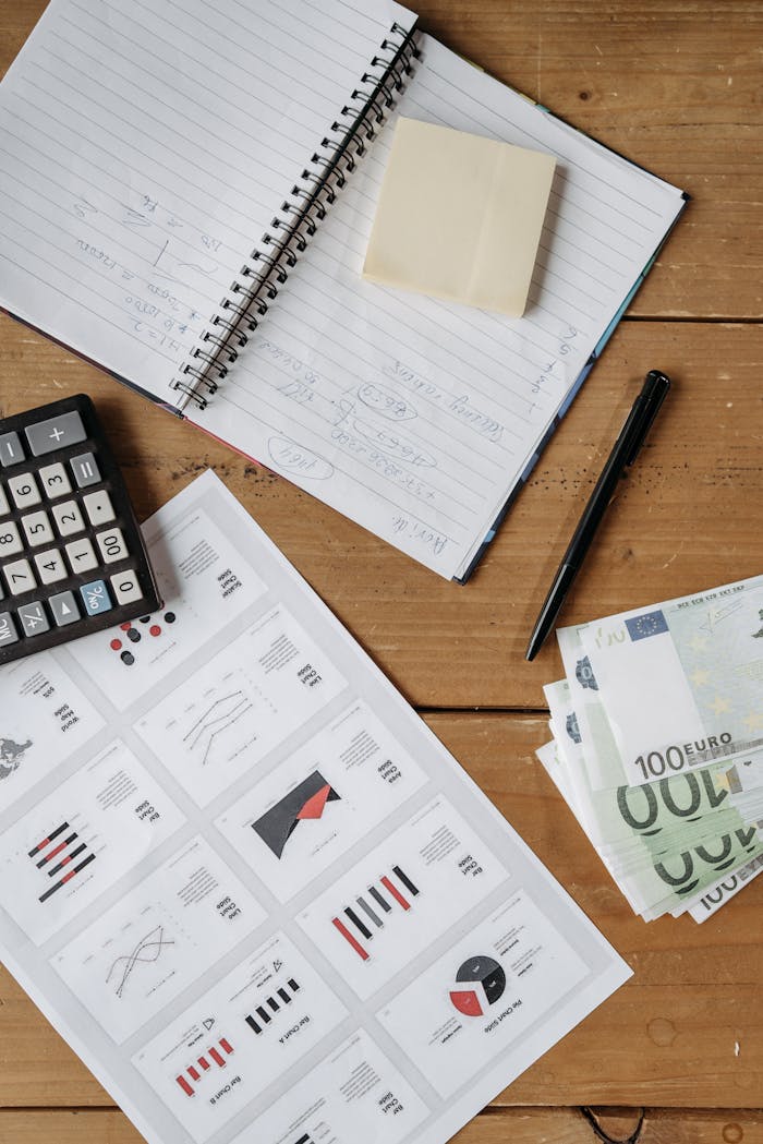 High-angle view of a financial workspace with charts, Euro bills, calculator, and notebook on a wooden desk.