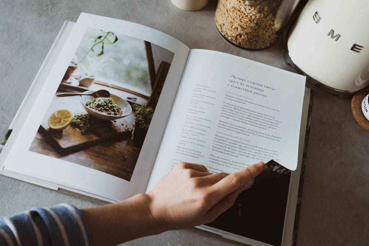 From above of crop faceless woman reviewing article in recipe book in kitchen